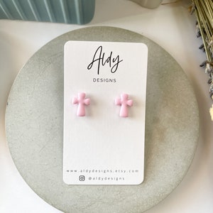 May include: A pair of pink cross-shaped stud earrings on a white card with the text "Aldy Designs" and the website address "www.aldydesigns.etsy.com" and the Instagram handle "@aldydesigns".