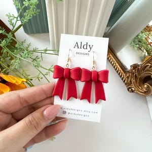 Könnte beinhalten: Rote Schleifenohrringe mit goldenen Haken. Die Ohrringe befinden sich auf einer weißen Karte mit dem Text "Aldy Designs" und Website-Informationen.