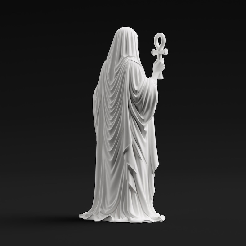 May include: A matte white statue of a cloaked figure viewed from behind, draped in flowing, detailed fabric that falls to the base. The figure holds an ankh symbol upright in one hand, with the entire sculpture presented against a solid black background.
