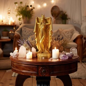 Peut inclure: Une statue dor&eacute;e repr&eacute;sentant une silhouette portant une coiffe en forme de croissant de lune et tenant un tissu drap&eacute; au-dessus de sa t&ecirc;te. La statue est entour&eacute;e de bougies blanches, de cristaux de quartz, d'am&eacute;thystes et de lavande s&eacute;ch&eacute;e.