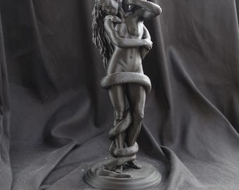 Puede incluir: Una escultura de color negro mate que representa una figura de cabello largo y fluido, de pie y entrelazada por una gran serpiente que se enrosca alrededor del torso y las piernas. La figura tiene los ojos cerrados y los brazos cerca de la cabeza.