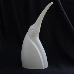 Op de afbeelding: Een matwit keramieken sculptuur die de gestileerde kop en nek van een vogel met een lange, slanke, omhoog wijzende snavel voorstelt. Het object heeft vloeiende lijnen en een gebogen silhouet, wat de essentie van een vogelvorm weergeeft.