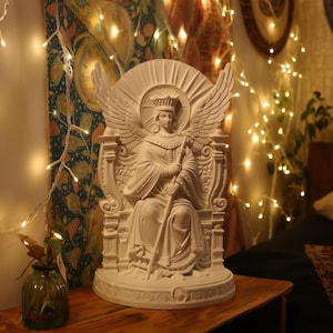 May include: A white statue of a seated angel with large wings, a crown, and a staff. The angel is seated on an ornate throne. The statue is on a wooden surface, with a vase and string lights in the background.