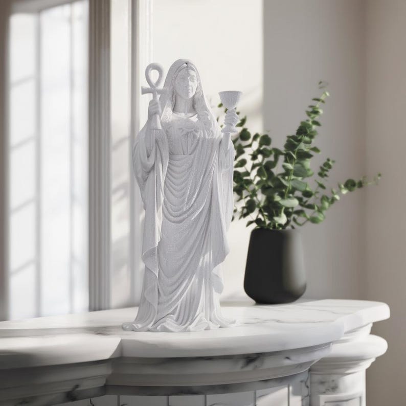 May include: A matte white statue of a standing figure draped in flowing robes. The figure holds an ankh symbol in the right hand and a chalice in the left hand. The sculpture features detailed folds in the fabric and a serene facial expression.