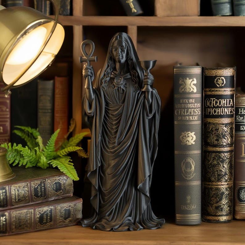 May include: A matte black figurine of a robed figure with long hair, holding an ankh symbol in the right hand and a chalice in the left hand. The figure wears a draped garment with a rose detail at the neckline and stands in a vertical pose.