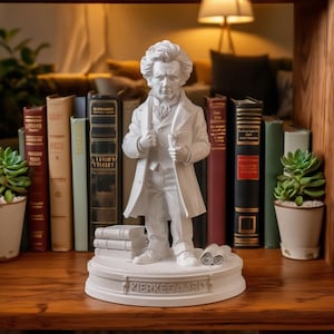 May include: A white statue of a man in a long coat, holding a small object, stands on a pedestal with the name "KIERKEGAARD" inscribed. The statue is surrounded by books and small potted plants, creating a scholarly aesthetic.