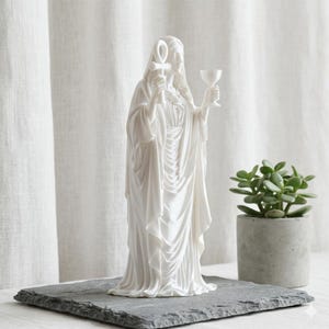 May include: A white matte statue of a robed figure standing upright. The figure holds an ankh symbol in the right hand and a chalice in the left hand. The sculpture features detailed flowing drapery and a hood covering the head, presented in a neutral tone.