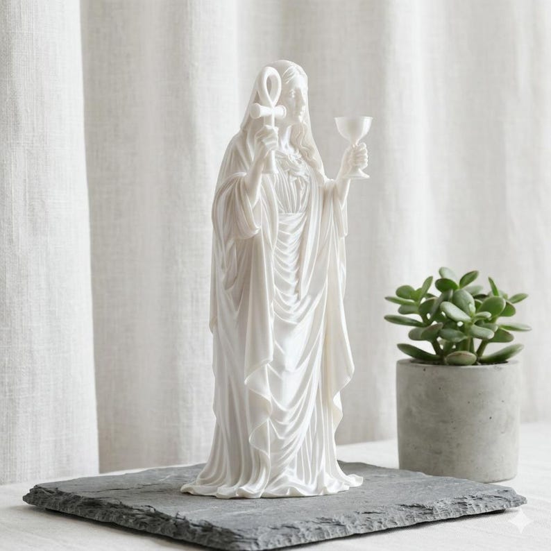 May include: A white matte statue of a robed figure standing upright. The figure holds an ankh symbol in the right hand and a chalice in the left hand. The sculpture features detailed flowing drapery and a hood covering the head, presented in a neutral tone.
