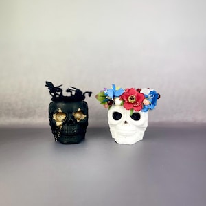 May include: Two decorative skull planters, one black with gold accents and the other white, both with floral crowns. The black skull planter has a gold glitter finish. The white skull planter has a red, blue, and white flower crown.