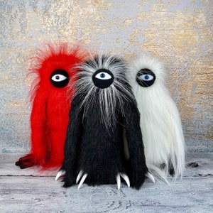 Può includere: Tre creature pelose con grandi occhi blu unici. La creatura a sinistra è rossa, quella al centro è nera e quella a destra è bianca.