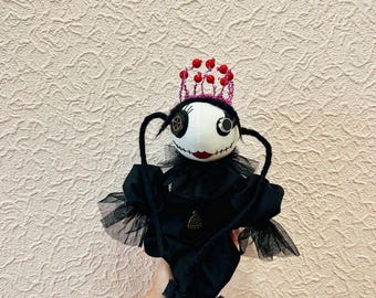 May include: A handcrafted fabric doll featuring a white face with stitched features, a black button eye, and a smaller eye detail. The doll wears a black ruffled dress with tulle sleeves, a red beaded crown, and has long black yarn hair.