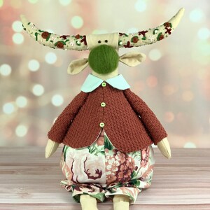 Puede incluir: Un juguete de peluche con forma de toro con cuernos marrones, una cara verde peluda, un suéter marrón y pantalones florales. El juguete está sentado sobre una superficie de madera.