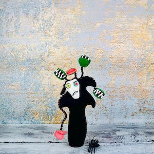 Peut inclure: Une figurine en feutre noir avec un visage blanc, des accents verts et rouges et une araignée. La figurine a trois formes de fleurs vertes et blanches et un chapeau rouge. Sur fond texturé blanc et or.