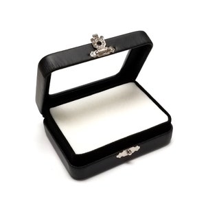 May include: Black rectangular jewelry box with a clear top and a silver-colored clasp. The interior is lined with white fabric. The box is open, revealing the inside. The box is made of a leather-like material.
