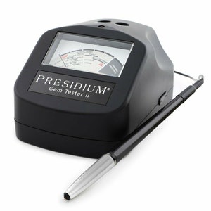 May include: A black Presidium Gem Tester II with a silver-tipped probe. The device has a digital display with a gauge for testing gemstones. The text "PRESIDIUM Gem Tester II" is printed on the front.