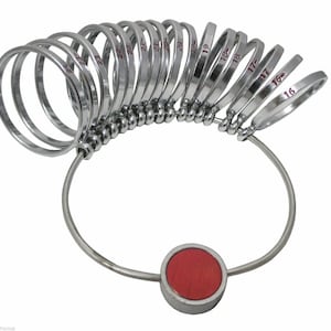 May include: A silver ring sizer with a red center piece. The sizer has a series of rings numbered 16 through 23.