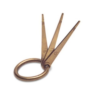 Peut inclure: Un ensemble de trois jauges de fil en laiton reliées par un anneau. Chaque outil a une extrémité pointue et est marqué d'un numéro, indiquant probablement la taille du fil. Les outils sont disposés en éventail, avec l'anneau à la base.