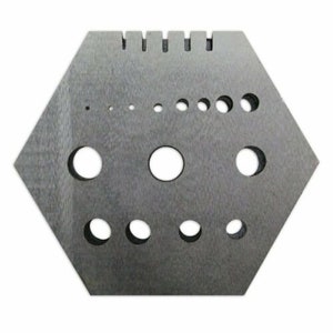 May include: A grey, hexagonal metal jewellery tool with various sized holes and notches. The tool has a textured surface and is designed for crafting or jewellery making. The holes are of different sizes, and the notches are at the top.