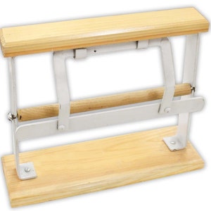 May include: A white metal paper towel holder with a wooden base and a wooden dowel. The holder is designed to hold a roll of paper towels and has a mechanism to tear off sheets.
