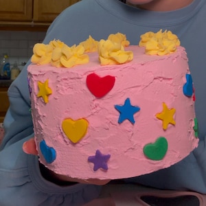 Puede incluir: Un pastel rosa decorado con glaseado amarillo, rojo, azul, verde y morado. El pastel está decorado con corazones y estrellas.