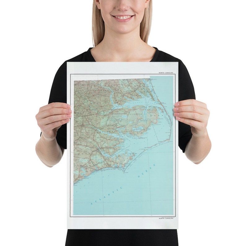 May include: A poster of a map of North Carolina, showing the state's coastline and major cities. The map is printed in shades of brown, green, and blue, with a light blue background. The poster is held by a person's hands.