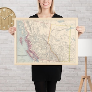 May include: Vintage map of British Columbia and Alberta, featuring detailed geographical features in muted tones of beige, pink, and green. The map is framed and displayed against a white brick wall, with a wooden clock and lamp in the background.
