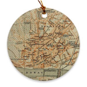 May include: A vintage map of the city of Vienna, Austria, printed on a round ceramic ornament. The map shows the city's streets, buildings, and landmarks, including the Danube River, the Hofburg Palace, and the Vienna State Opera. The map is in shades of brown, orange, and yellow.