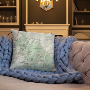 May include: Square pillow featuring a map of Catoctin Mountain Park, a National Park Service site. The pillow has a white background with green and red map details. The pillow is resting on a blue knitted blanket on a beige sofa.