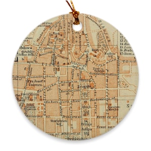 May include: A vintage map of Zagreb, Croatia, printed on a round ceramic ornament. The map shows streets, landmarks, and buildings in the city. The ornament is a light brown color with a gold string for hanging.