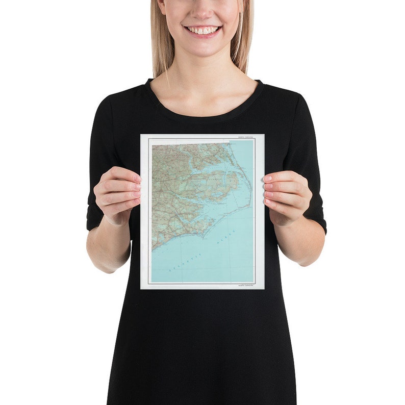 May include: A person holding a map print of the Outer Banks, North Carolina. The map is in shades of brown, green, and blue, and shows the coastline, roads, and waterways of the area.