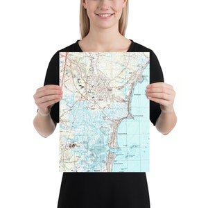 May include: A vintage-style map print of the coastal area of Wilmington, North Carolina. The map shows the city's streets, waterways, and surrounding areas. The map is printed in shades of blue, green, and brown.