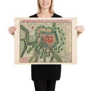 May include: A vintage map of Luxembourg City, titled "Nouveau Plan de la Ville de Luxembourg." The map is printed on paper with a cream background and features a detailed illustration of the city's layout, including its fortifications and surrounding landscape. The map is framed by a pink border and includes a key with explanations of the different features.