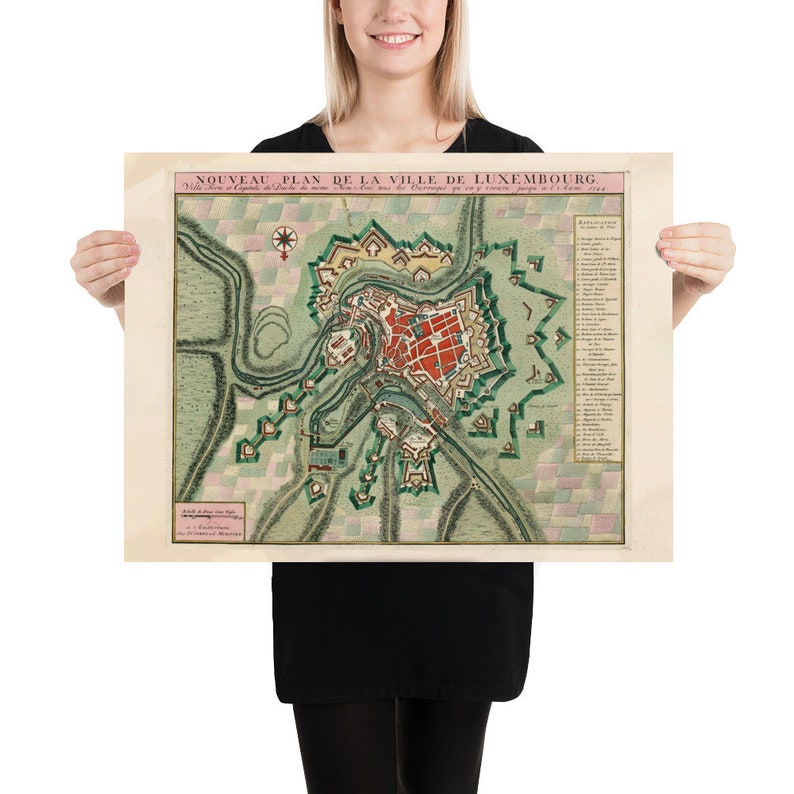 May include: A vintage map of Luxembourg City, titled "Nouveau Plan de la Ville de Luxembourg." The map is printed on paper with a cream background and features a detailed illustration of the city's layout, including its fortifications and surrounding landscape. The map is framed by a pink border and includes a key with explanations of the different features.