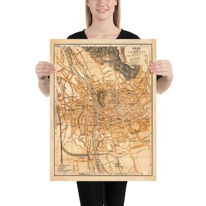 Könnte beinhalten: Vintage-Karte von Graz, Österreich, gedruckt in brauner und schwarzer Tinte auf cremefarbenem Papier. Die Karte zeigt die Straßen, Gebäude und Sehenswürdigkeiten der Stadt. Der Titel "GRAZ" ist in großen, fetten Buchstaben oben auf der Karte gedruckt.