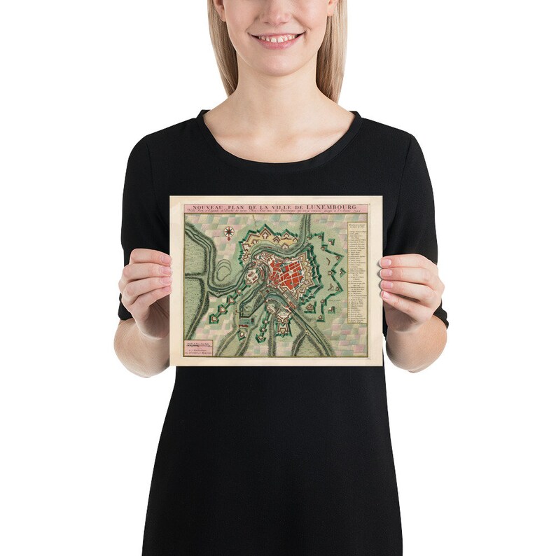 May include: A vintage map of the city of Luxembourg, printed in French. The map shows the city's fortifications and surrounding countryside. The map is in a light brown frame.