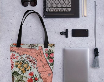 May include: A pink and white tote bag featuring a vintage map of Florida with floral illustrations. The map highlights cities and towns such as Jacksonville, Miami, and Tampa. The bag has black handles and a white background.