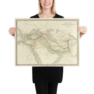 May include: A vintage map print of the Ancient Persian Empire, featuring detailed geographical information. The map is in muted tones of beige, green, and yellow, with the title "ANCIENT PERSIAN EMPIRE" at the top. The print is held by a person.