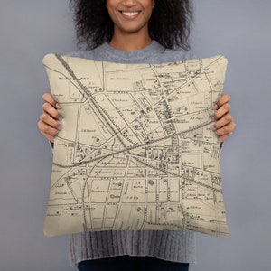 May include: A beige throw pillow featuring a vintage map of Durham, North Carolina printed on the front. The map depicts streets, buildings, and landmarks from the 19th century.