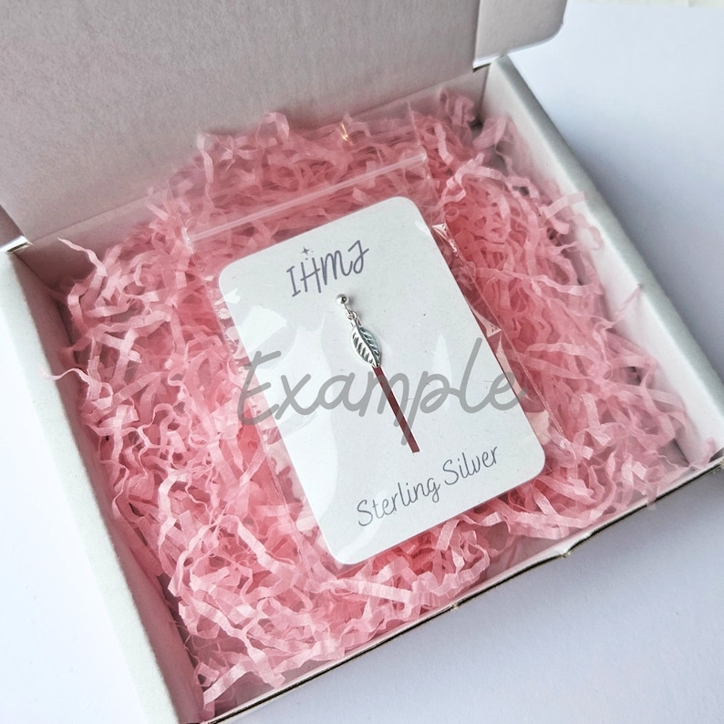 K&ouml;nnte beinhalten: Ein kleiner silberner Blattanh&auml;nger auf einer wei&szlig;en Karte mit dem Text "IHMz Example Sterling Silver". Die Karte befindet sich in einer durchsichtigen Plastikt&uuml;te und liegt auf rosa Seidenpapier in einer wei&szlig;en Box.