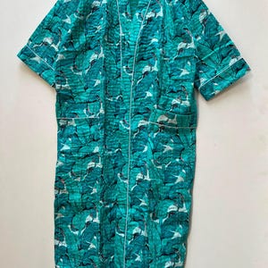 May include: A teal and white leaf-patterned robe with white trim. The robe has short sleeves and two front pockets. The robe is hanging on a wooden hanger against a white background. The robe is made of a quilted fabric.