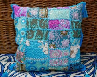 Puede incluir: Una funda de cojín de patchwork azul y rosa con detalles bordados y lentejuelas. La funda de cojín tiene un borla en cada esquina.