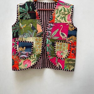 Puede incluir: Un chaleco patchwork con una variedad de coloridos estampados florales y de animales. El chaleco tiene un interior y un ribete a rayas negras y rosas, con dos bolsillos delanteros. El chaleco está colgado de una percha de madera.