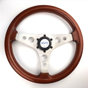 May include: A classic wooden steering wheel with a polished, dark brown finish. The wheel features a silver metal spoke design with three circular cutouts and a black center cap with the "NISMO" logo. The wheel is designed for a vehicle.