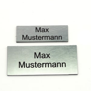 K&ouml;nnte beinhalten: Zwei silberne Namensschilder aus Metall mit dem Namen "Max Mustermann" in schwarzer Schrift.