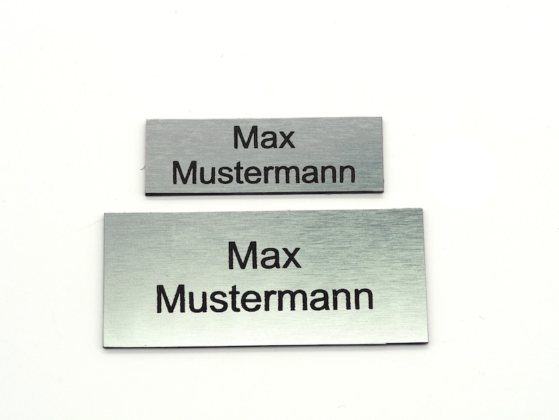 K&ouml;nnte beinhalten: Zwei silberne Namensschilder aus Metall mit dem Namen "Max Mustermann" in schwarzer Schrift.