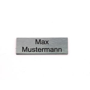K&ouml;nnte beinhalten: Ein silbernes Namensschild mit dem Text "Max Mustermann" in schwarzer Gravur.