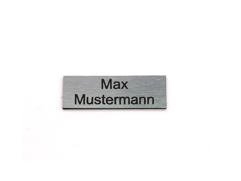K&ouml;nnte beinhalten: Ein silbernes Namensschild mit dem Text "Max Mustermann" in schwarzer Gravur.