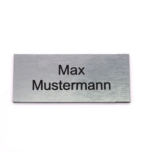 K&ouml;nnte beinhalten: Ein silbernes Namensschild mit dem in Schwarz gravierten Text "Max Mustermann".