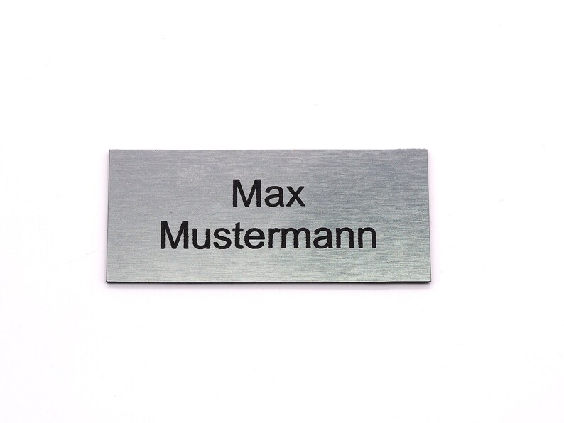 K&ouml;nnte beinhalten: Ein silbernes Namensschild mit dem in Schwarz gravierten Text "Max Mustermann".