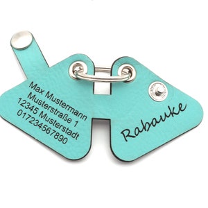 Könnte beinhalten: Ein türkisfarbener Leder-Schlüsselanhänger mit einem silbernen Ring und zwei silbernen Druckknöpfen. Der Schlüsselanhänger hat den Text "Max Mustermann Musterstraße 1 12345 Musterstadt 017234567890" und "Rabuake" eingraviert.